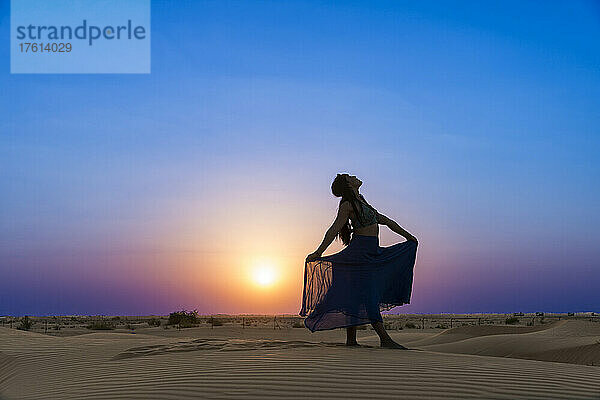 Frau mit langem Rock im Gegenlicht des Sonnenuntergangs im Wüstensand; Abu Dhabi  Vereinigte Arabische Emirate