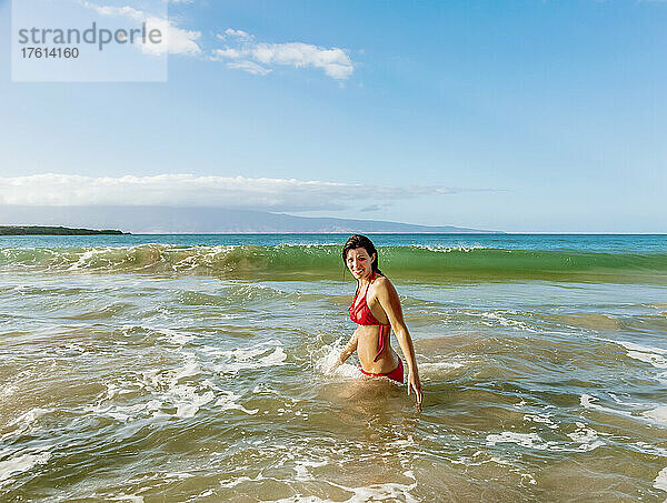 Frau im roten Bikini steht im Meerwasser vor der Küste und posiert für die Kamera am D. T. Fleming Beach mit der Insel Molokai in der Ferne; Kapalua  Maui  Hawaii  Vereinigte Staaten von Amerika