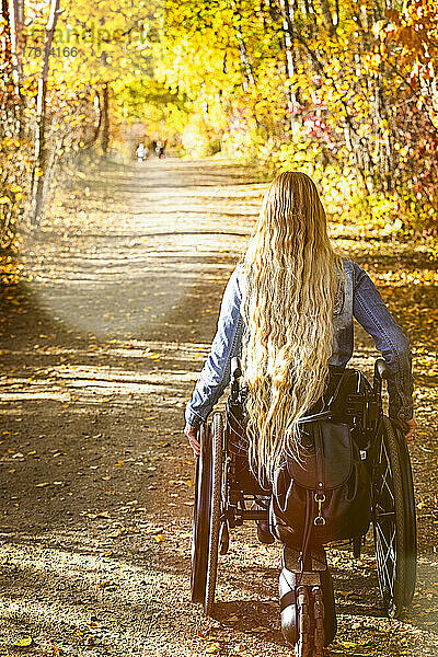 Junge querschnittsgelähmte Frau in ihrem Rollstuhl  die an einem schönen Herbsttag einen Weg in einem Park hinunterfährt; Edmonton  Alberta  Kanada