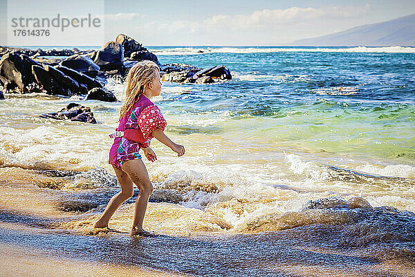 Ein junges Mädchen mit Schwimmflügeln  das am Strand der Kapalua Bay spielt und auf die Wellen wartet  mit der Insel Molokai im Hintergrund; Kapalua Beach  Maui  Hawaii  Vereinigte Staaten von Amerika