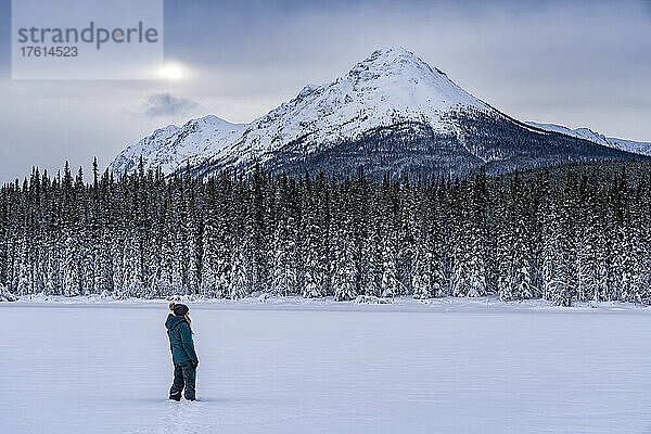 Frau  die im Schnee steht und die Aussicht betrachtet  während sie auf dem zugefrorenen Tagish Lake spazieren geht  während die Sonne durch die Wolken über den Berggipfeln lugt; Tagish  Yukon  Kanada