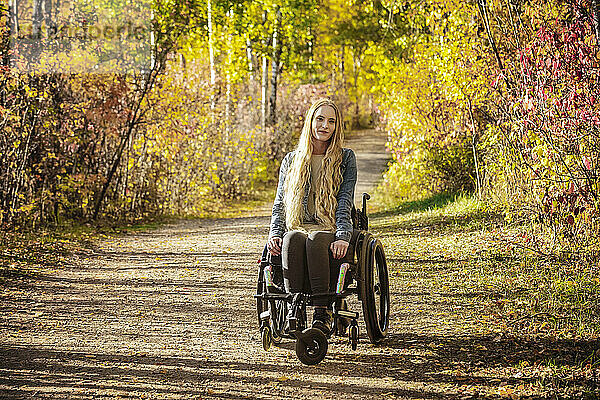 Junge querschnittsgelähmte Frau in ihrem Rollstuhl  die an einem schönen Herbsttag einen Weg in einem Park hinunterfährt; Edmonton  Alberta  Kanada