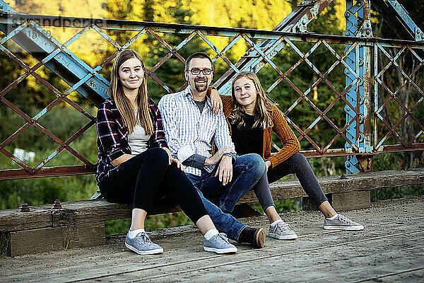 Außenporträt eines Vaters mit zwei Töchtern im Teenageralter  die im Herbst auf einer Brücke in einem Stadtpark sitzen; Edmonton  Alberta  Kanada