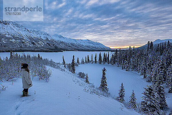 Frau steht im Schnee auf einem Berggipfel und blickt auf eine atemberaubende Landschaft mit der aufgehenden Sonne über dem Annie Lake im Winter; Whitehorse  Yukon  Kanada