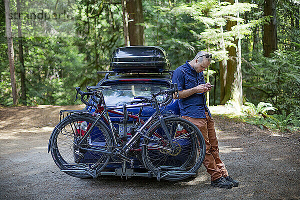 Mann steht an sein Auto gelehnt und benutzt sein Smartphone  mit Fahrrad auf dem Fahrradträger und Dachgepäckträger auf dem Auto; British Columbia  Kanada