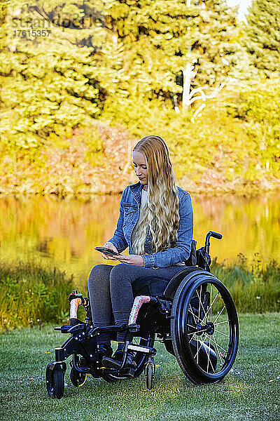 Junge querschnittsgelähmte Frau in ihrem Rollstuhl benutzt ein Smartphone in einem Park an einem schönen Herbsttag; Edmonton  Alberta  Kanada