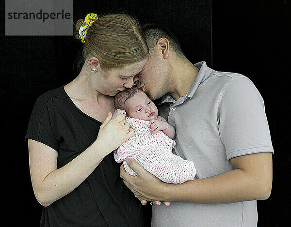 Familienporträt eines jungen Paares mit einem neugeborenen Mädchen vor einem schwarzen Hintergrund; Studio