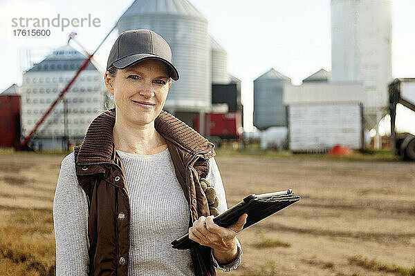 Porträt einer reifen Frau  die auf ihrer Farm arbeitet  einen Tablet-Computer in der Hand hält und vor einer Getreidemühle steht; Alcomdale  Alberta  Kanada