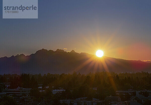 Brillanter Sonnenuntergang hinter den Bergen mit Sonnenstrahlen  die über den Wald und die Stadt darunter fluten; Surrey  British Columbia  Kanada