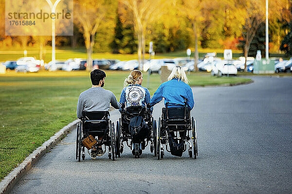 Drei junge Querschnittsgelähmte in ihren Rollstühlen  die an einem schönen Herbsttag über einen Parkplatz in einem Park fahren  sich an den Händen halten und sich gemeinsam in einer Reihe bewegen; Edmonton  Alberta  Kanada