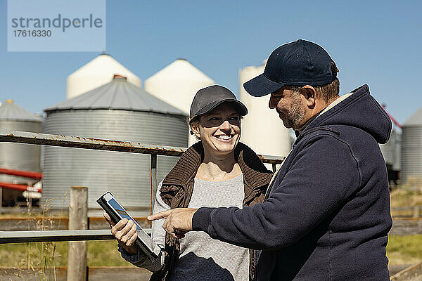 Älteres Paar  das sich anlächelt  während es an einem Zaun steht  mit Getreidespeichern im Hintergrund  und einen Tablet-Computer überprüft  um seine Farm zu verwalten; Alcomdale  Alberta  Kanada