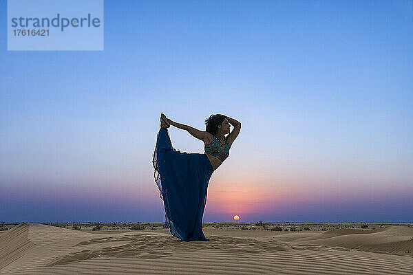 Frau mit langem Rock im Gegenlicht des Sonnenuntergangs auf dem Wüstensand  stehend in einer Tänzer-Yoga-Pose; Abu Dhabi  Vereinigte Arabische Emirate