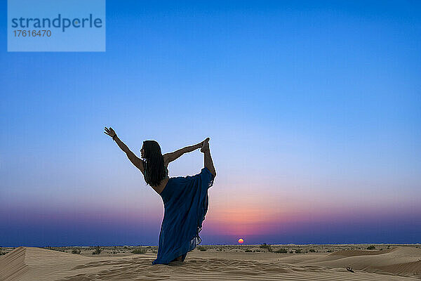 Frau mit langem Rock im Gegenlicht des Sonnenuntergangs auf dem Wüstensand  stehend in einer Tänzer-Yoga-Pose; Abu Dhabi  Vereinigte Arabische Emirate
