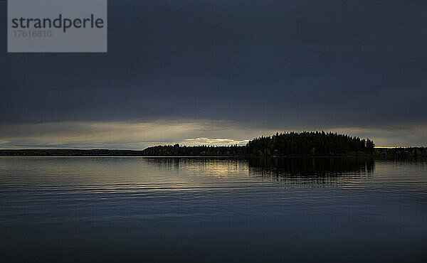 Silhouettierte Bäume entlang einer Uferlinie und das Licht des Sonnenuntergangs  das sich im ruhigen Wasser spiegelt  gesehen von der Meir Road im Lake Country  westlich von Prince George  BC  Kanada; British Columbia  Kanada