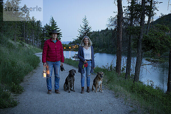 Ein Ehepaar geht in der Dämmerung mit seinen beiden Hunden auf einem Pfad um einen Teich spazieren; Kelowna  British Columbia  Kanada