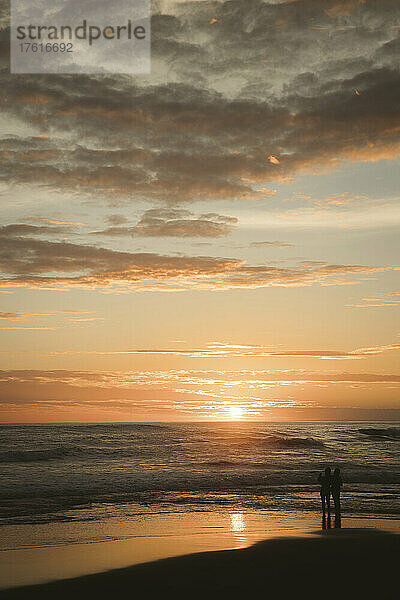 Silhouette von zwei Personen  die am Wasser stehen und den Sonnenuntergang am Strand beobachten; Munggu  Bali  Indonesien