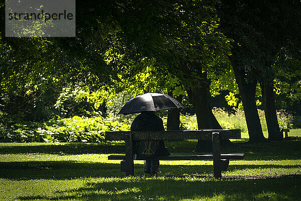 Person in schwarzer Kleidung unter einem schwarzen Regenschirm in einem Park an einem sonnigen Tag  die ihre Identität verbirgt; Hawthorn  Durham  England