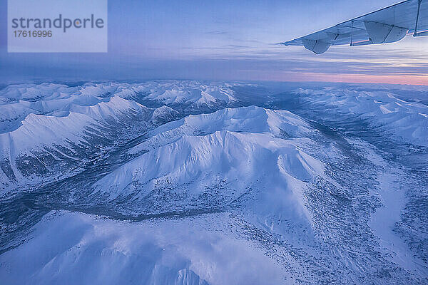 Luftaufnahme des Yukon im Winter mit einem Flugzeugflügel und einem rosafarbenen Sonnenaufgang über den schneebedeckten Bergen  die eine surreale Landschaft unter sich schaffen; Yukon  Kanada