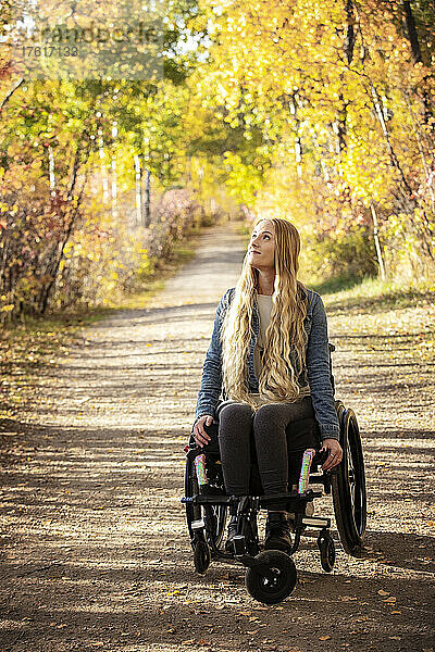 Junge querschnittsgelähmte Frau in ihrem Rollstuhl  die an einem schönen Herbsttag einen Weg in einem Park hinunterfährt; Edmonton  Alberta  Kanada