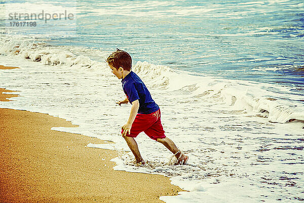 Energiegeladener Junge  der vor den Wellen am D. T. Fleming Strand am Rande des Wassers wegläuft; Kapalua  Maui  Hawaii  Vereinigte Staaten von Amerika