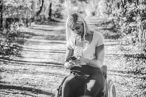 Junge querschnittsgelähmte Frau in ihrem Rollstuhl auf einem Weg in einem Park  die ihr Smartphone benutzt; Edmonton  Alberta  Kanada