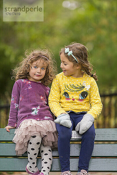Porträt von zwei jungen Schwestern  die zusammen auf dem oberen Rand einer Parkbank sitzen  wobei die eine in die Kamera schaut und die andere ihre Schwester beobachtet; Toronto  Ontario  Kanada