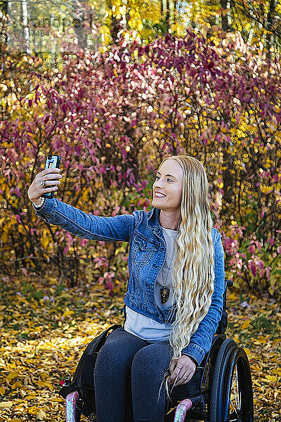 Eine junge querschnittsgelähmte Frau in ihrem Rollstuhl macht ein Selbstporträt mit ihrem Smartphone in einem Park an einem schönen Herbsttag; Edmonton  Alberta  Kanada