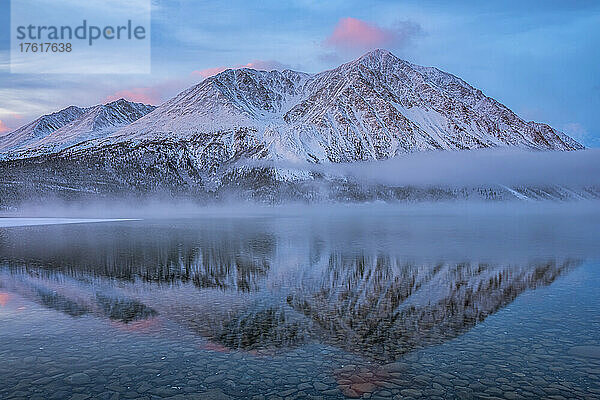 Winterliche Landschaft mit schneebedeckten Bergen  die sich im stillen Wasser des Kathleen Lake spiegeln  mit nebligen  tief hängenden Wolken und Pastellfarben  die eine stimmungsvolle Atmosphäre schaffen; Haines Junction  Yukon  Kanada