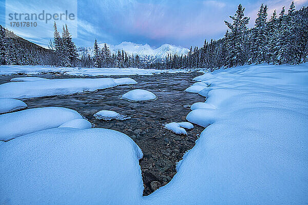 Schneebedeckte  eisumrandete Felsen am Wheaton River  mit rosa Wolken über den Bergen in der Ferne; Whitehorse  Yukon  Kanada