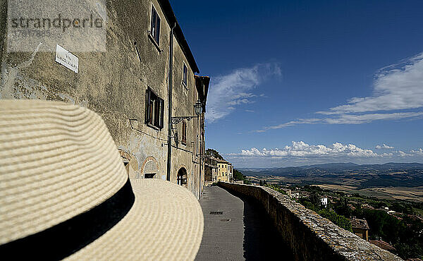 Nahaufnahme des Strohhuts eines Touristen von hinten  der die von Steinmauern gesäumte Gasse mit Blick auf die Landschaft in der historischen Altstadt von Volterra hinunterschaut; Volterra  Toskana  Italien