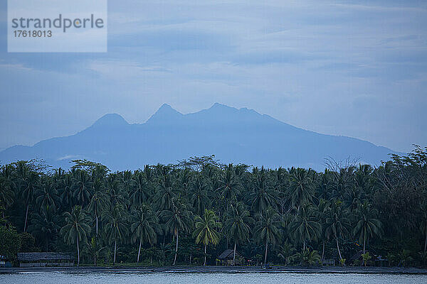 Dschungel und Küstendorf zwischen Kokospalmen (Cocos nucifera) mit Silhouetten der Berge in der Ferne an der Küste der Provinz Morobe; Provinz Morobe  Papua-Neuguinea