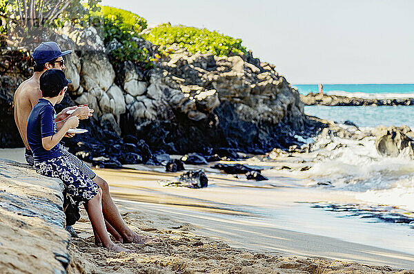 Ein Vater und sein Sohn sitzen an einem großen Felsen am Strand  essen und verbringen Zeit miteinander und blicken auf den Ozean in der Kapalua Bay; Kapalua  Maui  Hawaii  Vereinigte Staaten von Amerika