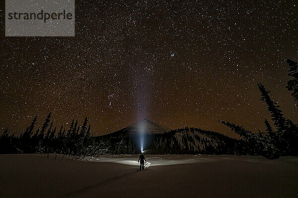 Blick von hinten auf eine Frau  die in den Nachthimmel starrt  während sie mit einer Stirnlampe durch eine schneebedeckte Winterlandschaft mit funkelnden Sternen am Himmel geht; Whitehorse  Yukon  Kanada