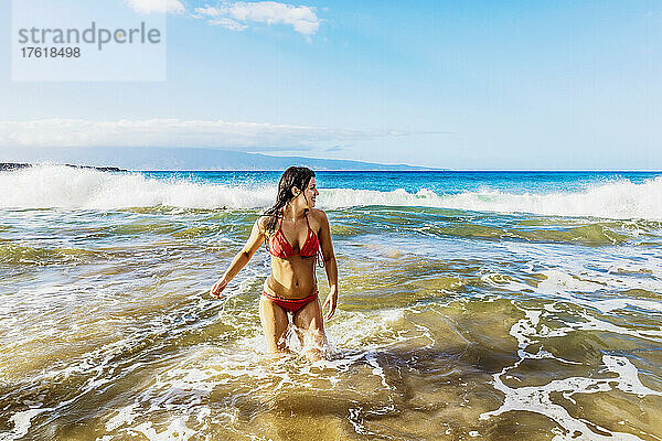 Frau im roten Bikini steht im Meerwasser vor der Küste und spielt in den Wellen am D. T. Fleming Beach mit der Insel Molokai in der Ferne; Kapalua  Maui  Hawaii  Vereinigte Staaten von Amerika