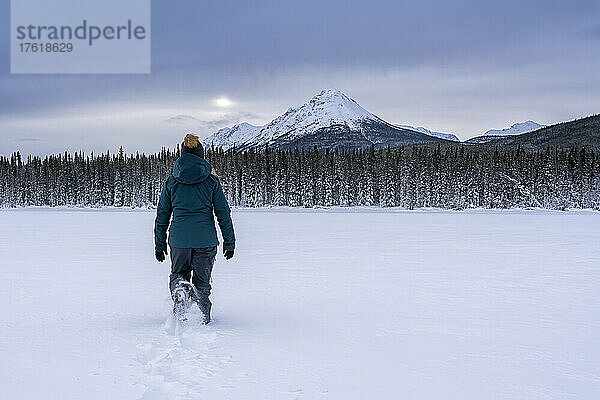 Blick von hinten auf eine Frau im Schnee  die auf dem zugefrorenen Tagish Lake spazieren geht  während die Sonne durch die Wolken über den Berggipfeln lugt; Tagish  Yukon  Kanada