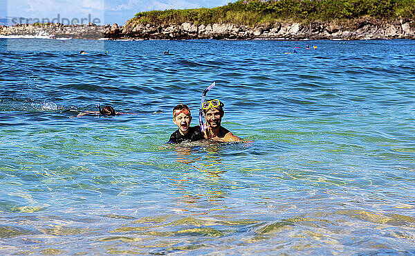 Ein Vater und sein kleiner Sohn im Meerwasser der Kapalua Bay mit Schnorchelausrüstung und posieren für die Kamera  während ein anderer Sohn im Hintergrund weiter schnorchelt; Kapalua  Maui  Hawaii  Vereinigte Staaten von Amerika