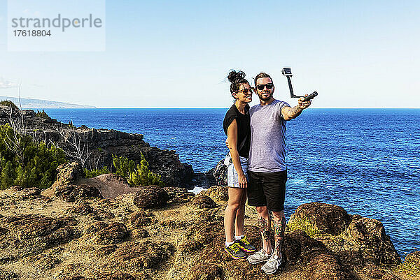 Ein junges Paar steht am Ufer von Nakalele Point und macht ein Selbstporträt mit der Küstenlinie der Insel und der Insel Molokai im Hintergrund; Maui  Hawaii  Vereinigte Staaten von Amerika