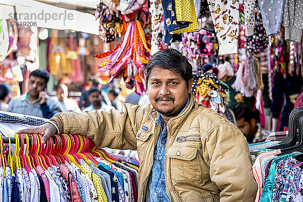 Ein Mann steht in einem Bekleidungsstand auf einem Markt und stützt sich auf ein Gestell mit farbenfrohen Kleidungsstücken  die zur Schau gestellt werden; Dehli  Indien