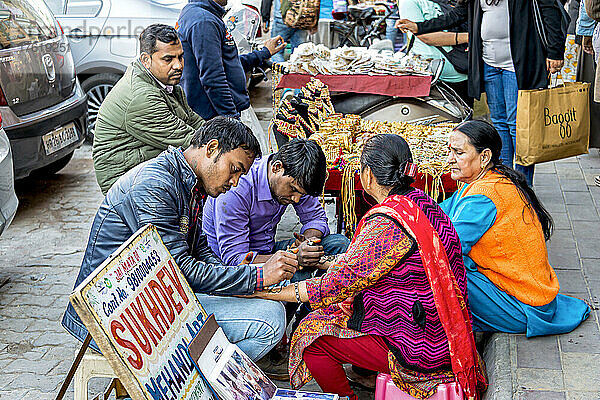 Frau  die sich mit Henna-Tattoos (Mehndi-Kunst) die Hände tätowieren lässt  während sie auf einer Straße neben einem Schmuckhändler sitzt; Amritsar  Punjab  Indien