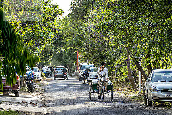 Straßenszene in einem Dorf in Indien mit Fahrzeugen und Motorrädern sowie einem Mann  der mit einem Anhänger fährt und telefoniert; Greater Noida  Uttar Pradesh  Indien