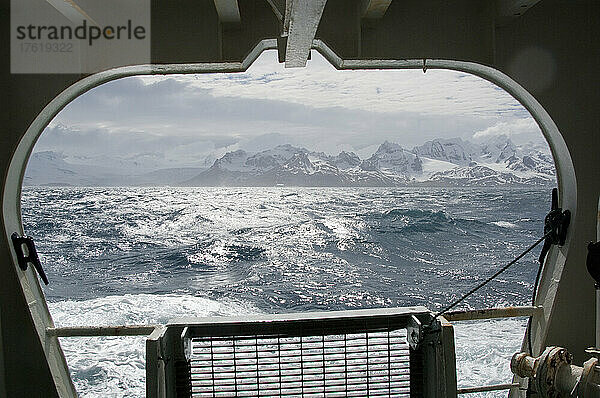 Blick aus dem Fenster eines Schiffes auf den Wellengang des Südlichen Ozeans und die schneebedeckten Berge der Insel Südgeorgien an einem sonnigen Tag; Südgeorgien  Antarktis