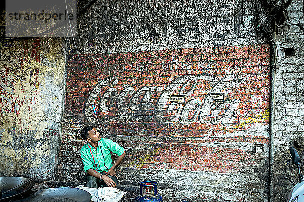 Mann sitzt und schaut auf ein verblasstes Cola-Schild  das auf eine Backsteinmauer gemalt ist; Amritsar  Punjab  Indien