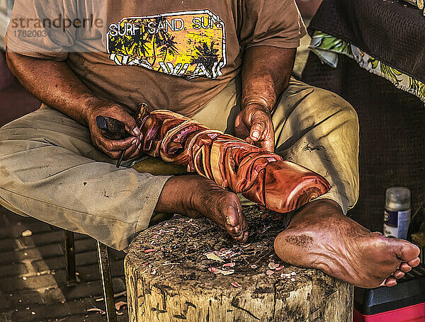 Ein hawaiianischer Mann sitzt barfuß und schnitzt mit einem Werkzeug Holz  um ein traditionelles Kunsthandwerk herzustellen; Maui  Hawaii  Vereinigte Staaten von Amerika