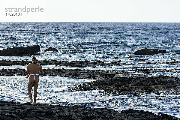 Ein Mann in einem Malo (Lendenschurz) steht am Ufer des Wassers. Er ist der Parkführer  Kulturdemonstrator und Dolmetscher im Pu?uhonua o Honaunau National Historical Park; Honaunau  Insel Hawaii  Hawaii  Vereinigte Staaten von Amerika