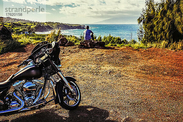 Ein Mann sitzt auf einem Felsen und blickt auf den Ozean mit der Insel Molokai in der Ferne und seinem Motorrad  das hinter ihm an einem Aussichtspunkt am Honoapiilani Highway geparkt ist; Maui  Hawaii  Vereinigte Staaten von Amerika