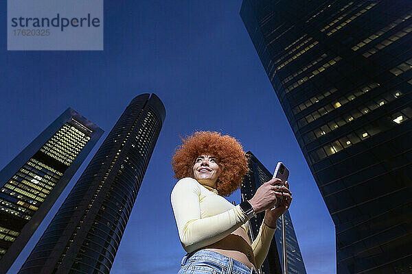 Lächelnde Frau mit Afro-Frisur mit Smartphone vor Wolkenkratzern