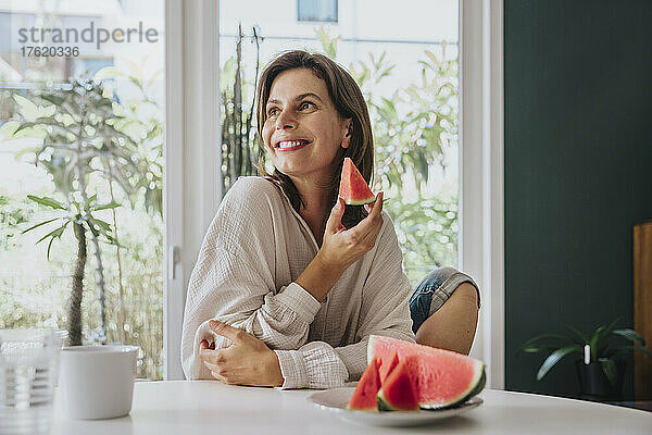 Glückliche Frau hält eine Wassermelonenscheibe in der Hand und sitzt zu Hause