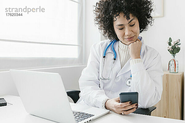 Arzt sitzt mit Laptop und benutzt Mobiltelefon in der Klinik