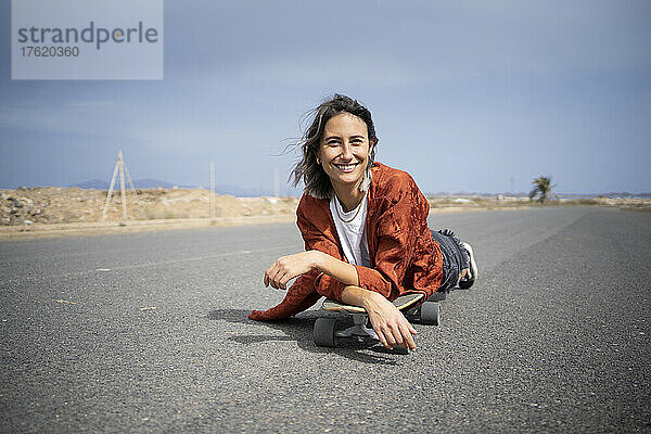 Lächelnde junge Frau liegt auf einem Skateboard auf der Straße