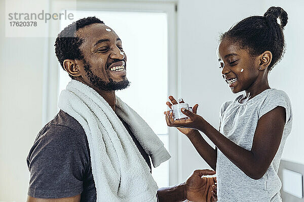 Lächelnde Tochter trägt im Badezimmer Feuchtigkeitscreme auf den Vater auf